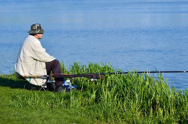 Pescador sentado en la hierba verde cerca del lago y la pesca —  Fotos de Stock