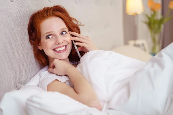 Улыбающаяся рыжая женщина разговаривает по мобильному телефону в постели — стоковое фото