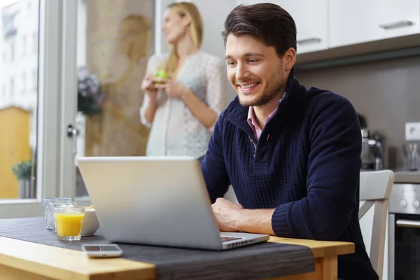 Rindo homem usando laptop com esposa no fundo — Fotografia de Stock