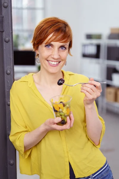 Niedliche rothaarige Frau nascht bei der Arbeit an Obst — Stockfoto