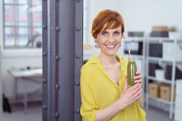 Счастливая женщина с зеленым напитком здоровья на работе — стоковое фото