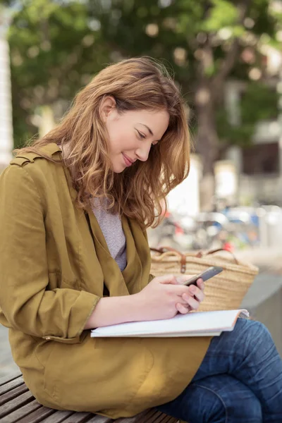 Lächelndes Mädchen auf der Bank, das auf ihr Handy schaut — Stockfoto