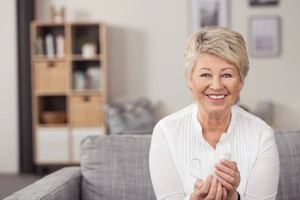 Mujer de mediana edad feliz sosteniendo una taza en el sofá —  Fotos de Stock
