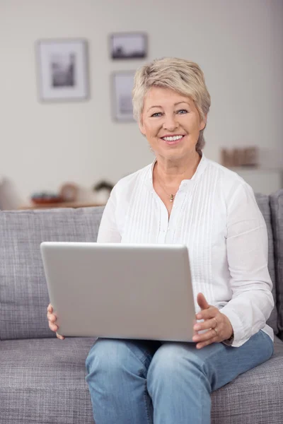 Femme blonde senior souriante avec ordinateur portable au canapé — Photo