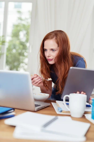 Mujer en su oficina leyendo en el ordenador portátil mientras come — Foto de Stock