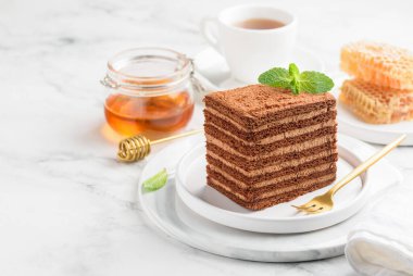 Mermer bir masada beyaz bir tabakta bir dilim çikolatalı bal pastası. Bir fincan çay, bal peteği, bir kavanoz bal. Seçici odaklanma. Boşluğu kopyala.