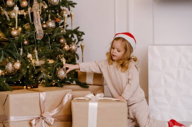 Noel ağacının altında Noel hediyesi olan Noel Baba şapkalı bir kız. Evde Noel hediyesi olan bir çocuk var. Kış tatili için dekore edilmiş. Yumuşak odak