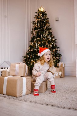 Noel Baba şapkalı, Noel ağacı arkaplanlı bir kız. Evde Noel hediyesi olan bir çocuk var. Kış tatili için dekore edilmiş. Yumuşak odak