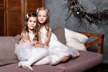 Beyaz elbiseli iki tatlı kız kardeş birbirlerine yakın koltukta oturup evde oynuyorlar. Küçük kız kardeşi en küçük kardeşine sarılıyor. Evde çocuklu bir aile. Boşluğu kopyala.