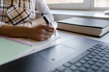 Genç bir kadın, öğrenci ev ofisinde uzun mesafe eğitim kursu alıyor ve dizüstü bilgisayarla çevrimiçi kurs çalışırken kağıt defterine kalemle içerik yazıyor. Kapat..