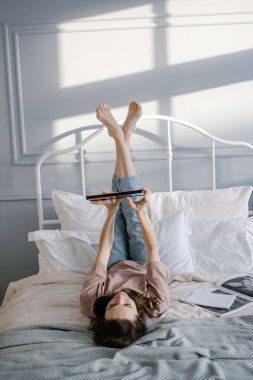 Yaratıcı kadın tabletle yatakta yatıyor, düşünceli bir şekilde gülümsüyor, bir mesaj yazıyor, görüntülü konuşma yoluyla arıyor, haberleri okuyor. Evden çalışma, serbest çalışma ve yaşam tarzı konsepti. 