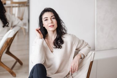 Zarif giysiler içinde genç, gülümseyen bayan girişimci çağdaş iş yerinde oturuyor ve kameraya bakıyor.