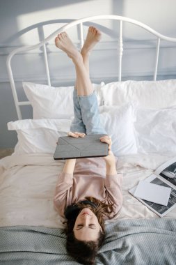 Yaratıcı kadın tabletle yatakta yatıyor, düşünceli bir şekilde gülümsüyor, bir mesaj yazıyor, görüntülü konuşma yoluyla arıyor, haberleri okuyor. Evden çalışma, serbest çalışma ve yaşam tarzı konsepti. 