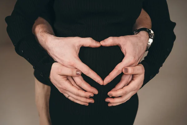 Ένας ευτυχισμένος άντρας στέκεται δίπλα στην έγκυο κοπέλα του. Ένας άντρας δείχνει μια καρδιά σε μια έγκυο σύζυγο από τα δάχτυλα των χεριών του. Ένας άντρας χαίρεται που γίνεται πατέρας. — Φωτογραφία Αρχείου