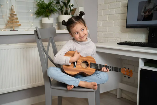 En liten flicka spelar ukulele. Kreativ utveckling hos barn. Musikutbildning från barndomen. Undervisning musik på nätet hemma — Stockfoto