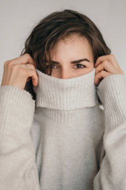 Güzel bir esmerin dikey çerçevesi. Gri kazaklı bir kızın samimi bir fotoğrafı. Kız yüzünü bir ceketle örtüyor. Bir kızın gözlerini kapat.