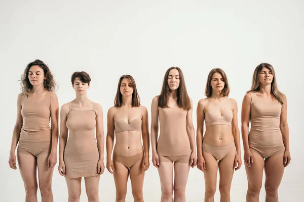 Several girls with different physiques. Women in beige underwear. Different height and weight in women. The girls stand in one row and look into the frame