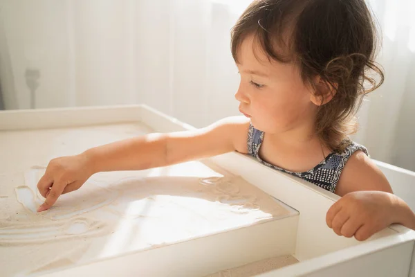 Mesa de dibujo de arena para niños. Una niña dibuja con el dedo sobre una mesa de arena clara. Desarrollo creativo en niños pequeños. —  Fotos de Stock