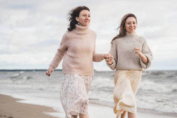 Two girls hold hands and run along the seashore. The sisters have fun together. A couple in love on a date. Cozy autumn walk along the sea