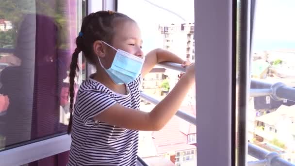 Une petite fille est tombée malade en vacances. La fille manque la maison à cause de la maladie. Des enfants avec un masque médical. Une fille regarde par la fenêtre pendant que d'autres enfants marchent. — Video