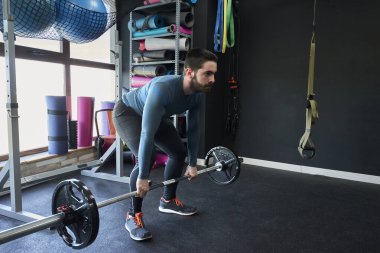 Spor kıyafetli genç adam spor salonunda ağırlık kaldırırken gülümsüyor.