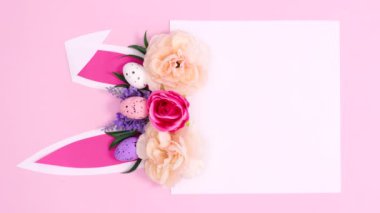 Tavşan kulaklı, dikey yaratıcı kağıt kart notu ve pastel pembe arka planda çiçek açan bahar düzenlemesi. Paskalya düzlüğü stop hareketi kompozisyonu