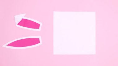Tavşan kulaklı ve çiçekli dikey kağıt kart notu pastel pembe arka planda görünür. Paskalya düzlüğü stop hareketi kompozisyonu