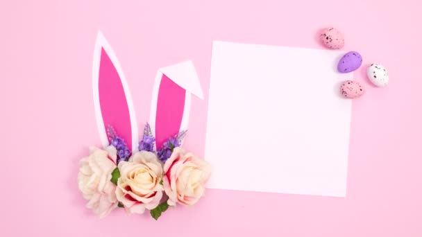 パステルピンクの背景に紙のカードノートの横に花の配置でウサギの耳が移動します イースターフラットレイストップモーション — ストック動画