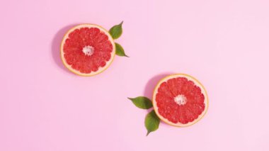 Two sliced fresh grapefruits with green leaves rotating on pastel pink background. Stop motion flat lay