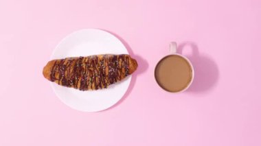 Pastel pembe arka planda bir fincan kahveyle dönen taze, lezzetli çikolatalı kruvasan. Hareketi durdur
