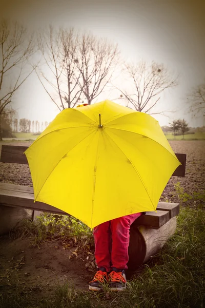 Boy sitting on a bench hidden by a yellow umbrella — Stock Photo, Image