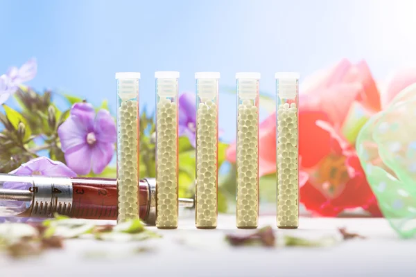 Tubos de vidrio pequeños con glóbulos homeopáticos, jeringa y flores — Foto de Stock