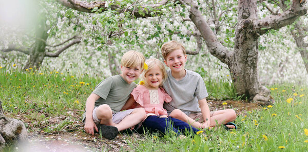 Three happy children, brothers and their sister are smiling happily as they sit outside under the flowering apple trees on a spring day.
