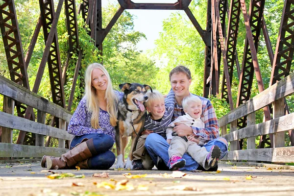 Porträt einer glücklichen Familie und eines Hundes, die im Herbst auf einer Brücke sitzen — Stockfoto