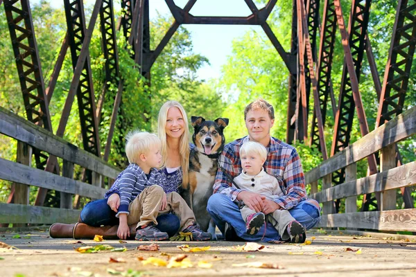 Familia de cuatro personas y perro sentados en el puente en otoño — Foto de Stock