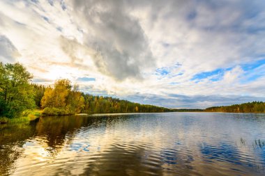 Orman gölünün üzerindeki bulutlu gökyüzünde gün batımı. Kıyıdan görünüm