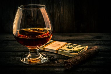 Glass of brandy with pack of dollars and cuban cigar on an old wooden table