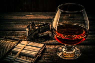 Glass of brandy and cigarette case with cigarettes and lighter on an old wooden table 