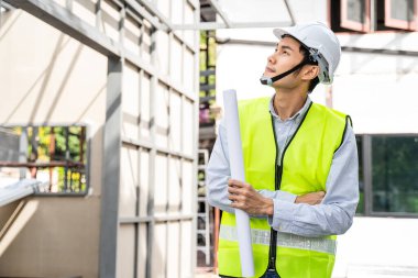 Asyalı işçi, mimari yapı ve inşaat alanındaki çalışma sürecini kontrol ediyor. İnşaat tarafında çizim yapan erkek mühendisler kendinden emin bir yüzle çalışmalarını izliyorlar..