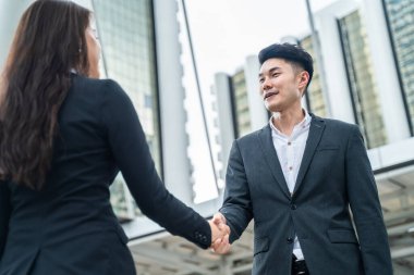 Asyalı işkadını ve erkek arka plandaki belediye binasında el sıkışırlar. Pazarlık tamamlandıktan sonra bir ortaklık anlaşması başarılı olur. İş anlaşması, birleşme ve satın alma kavramları.