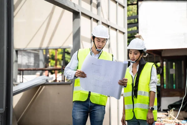 Asian Colleagues worker Specialists team wearing protective safety helmet look at blueprint on Construction Site. Civil Engineer Male and architect discuss about Real Estate Building Project work plan