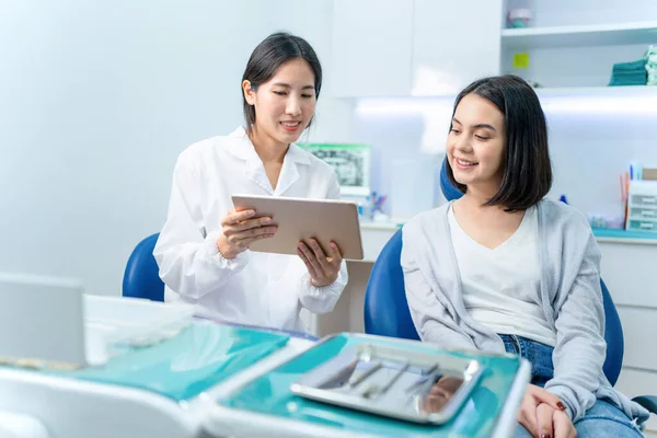 Asyalı genç bir dişçi dijital tablet tutuyor ve beyaz bir genç kıza danışmanlık yapıyor. Diş sağlığı kliniğinde sunulan oral tedavi hizmetini açıklayan doktor.