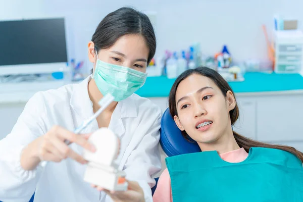 Asyalı genç bir kız diş hekimi, diş bakımı ve diş fırçalama konusunda ortodontisi olan insanlara açıklama yapıyor. COVID-19 'u önlemek için diş kliniğinde koruyucu yüz maskesi takan ortodontist..