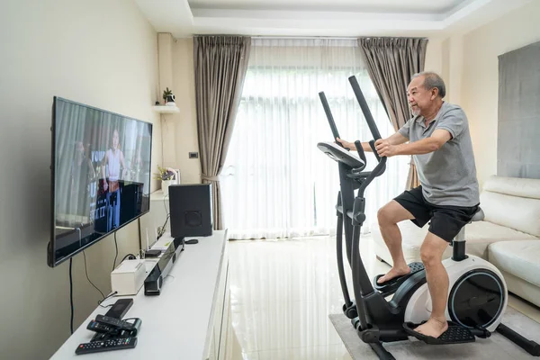 Asyalı yaşlı adam evde bisiklet egzersiz makinesi üzerinde egzersiz yapıyor. İyilik için. Aktif yaşlı erkek tecrit aktivitesi yapıyor. Evdeki spor eğitmeninden alınan talimatları takip ediyor.