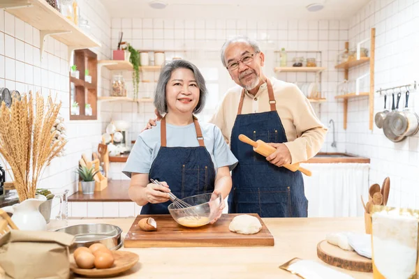 Portrait of Asian happy Senior elderly couple standing in kitchen at house feel happy and enjoy family retirement life together. Loving Older grandparents with apron, smiling and looking at camera.