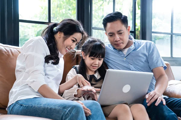 Asian happy family, young little girl kid with parents spend time together using laptop playing internet online in living room at home. Father and Mother teaching daughter from video virtual learning.