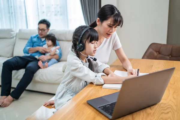 Asian happy family stay home, parents spend time with young little kid daughter in living room together. Loving mother teach and support small girl use computer for virtual learning and doing homework