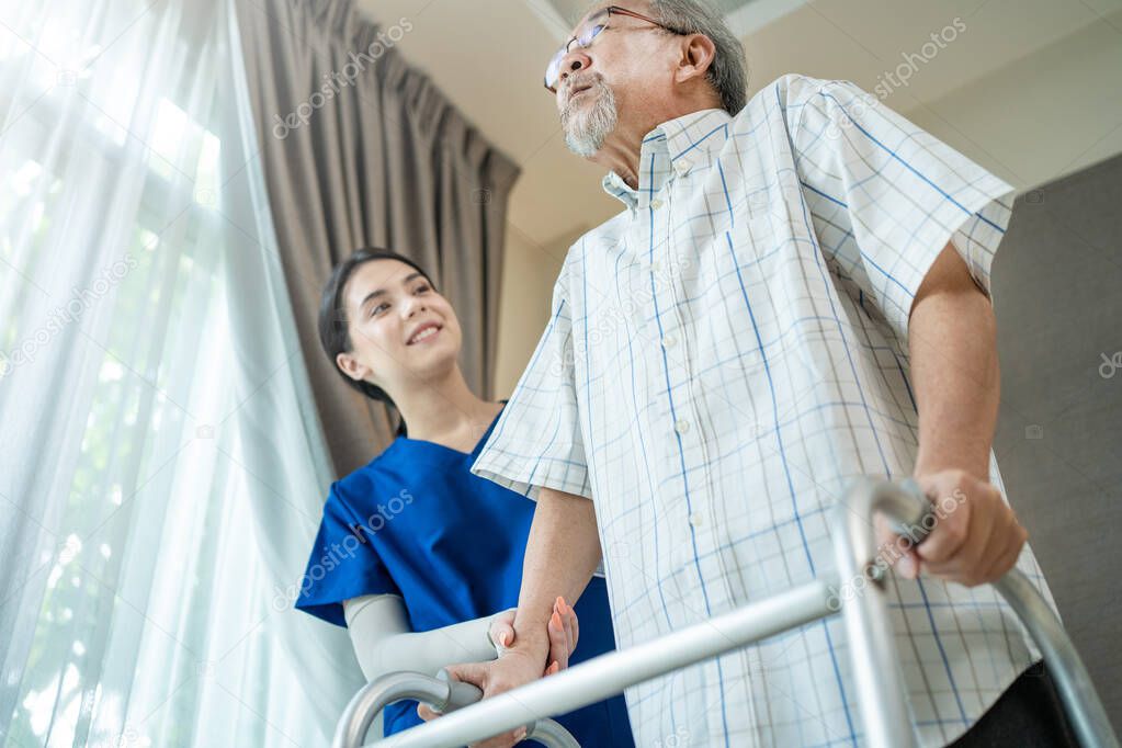 Asian Senior elderly disabled man patient walking slowly with walker at nursing home care. Caregiver therapist nurse support older handicapped male use Zimmer frame. Medical therapy insurance concept.