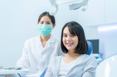 Diş hekiminin ya da maske takan bir doktorun portresi genç güzel bir kızın arkasında oturuyor dişleri beyazlıyor gibi gülümsüyor ve diş sağlığı kliniğinde tedavi sonrası kameraya bakıyor.
