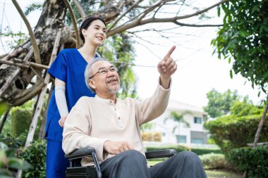 Asyalı mutlu emekli büyükbaba, yeşil parkı ve genç kadın hemşireyi göstererek dışarıda eğleniyor. Bakıcı doktor tekerlekli sandalyedeki yaşlı hastayı gülümseyerek itiyor, şömine bakımının keyfini çıkarıyor..
